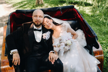 The bride and groom with a bouquet are sitting in a carriage in nature in retro style