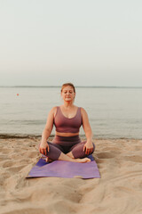 Fototapeta na wymiar Caucasian woman practicing yoga at seashore sandy beach on sunrise. Womens health and wellness. Sports body positive