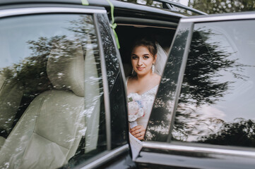 A young, smiling, beautiful bride in a white dress with a bouquet in her hands sits in a black car with an open door and window. Wedding portrait, photography.
