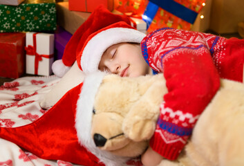 Child girl sleeping in new year or christmas decoration. Holiday lights and gifts, Christmas tree decorated with toys. She's wearing a red sweater and a Santa helper hat