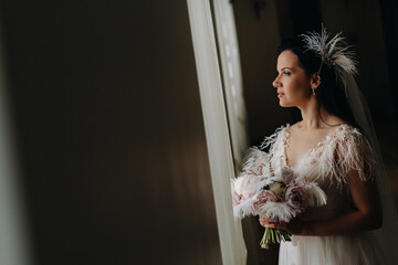 The bride in a wedding dress and with a bouquet stands at the old window and looks