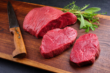 Raw dry aged bison beef rump steak piece and slices offered with herbs as close-up on wooden design board