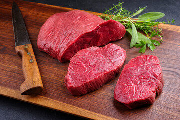 Raw dry aged bison beef rump steak piece and slices offered with herbs as close-up on wooden design...