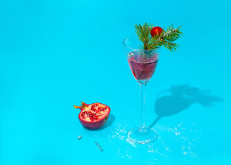 Modern cocktail glass with an exotic drink with small bubbles decorated with fresh green pine branches. Red decorative Christmas ball with pomegranate and glass shadow on a turquoise background.