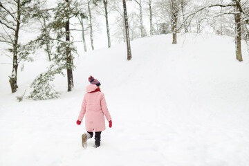 Fototapeta premium Adorable girl having fun in beautiful winter park during snowfall. Cute child playing in a snow. Winter activities for family with kids.