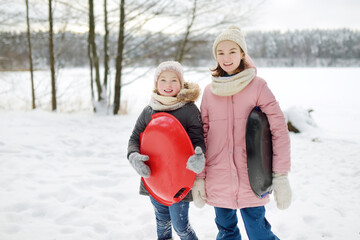 Two funny young girls having fun with a sleigh in beautiful winter park. Cute children playing in a snow.