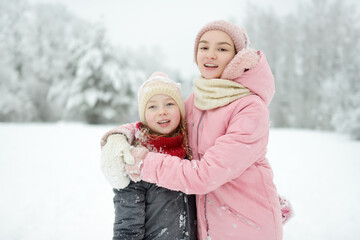 Fototapeta premium Two young girls having fun together in beautiful winter park. Cute sisters playing in a snow. Winter activities for family with kids.