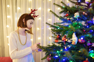 Adorable girl decorating the Christmas tree with colorful glass baubles. Trimming the Christmas tree. Celebrating Xmas at home.