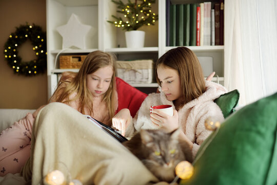 Two young sisters snuggling up on the sofa in a cozy living room at Christmas. Cute children using a tablet at home during winter break. Kids reading a book in comfy blanket.