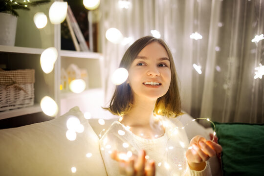 Beautiful teenage girl playing with Chistmas lights in a cozy living room on Christmas eve. Celebrating Xmas at home.