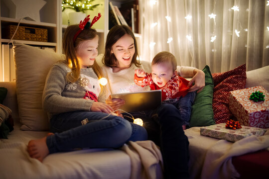 Two girls and a baby boy using a tablet pc at home in warm and cozy living room at Christmas. Family having online video call on Xmas eve.
