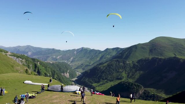 Paragliders take off from the side of a mountain, while others are setting up their equipment. Spectators watching.