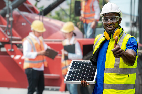 Factory engineer African man standing confidence and holding solar cell panel for renewable energy engineering. workers inspecting and meeting in front of heavy machine vehicle car.