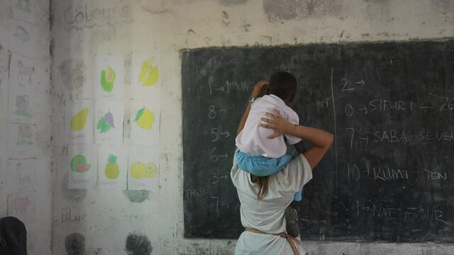 White caucasian teacher with black African child lifted on her shoulders helping him to write on the blackboard at school. Teaching numbers in Swahili. Beautiful volunteer cinematic shot. Africa 4K.