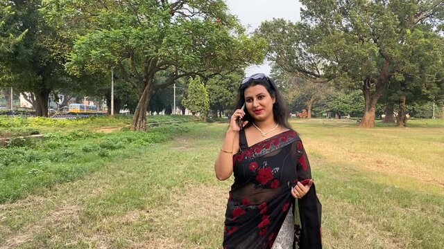 A young beautiful woman in black saree is smiling and talking on a mobile outdoor in a park. A happy and attractive Indian girl is on a phone and walking in park.
