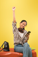 happy girl with raised fist up and looking at camera while holding and using cellphone while sitting