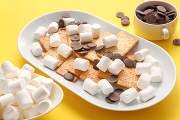 Plate with crackers, marshmallows and chocolate on yellow background, closeup