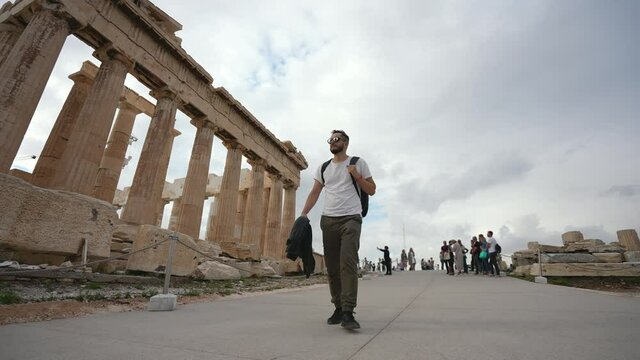 Young adult tourist among crowd of people, walking, looking around, mesmerized by ancient Greek architecture. Traveler on sight of Acropolis in Athens. Tourism, traveling to ruins of old civilization.