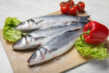 Board with fresh uncooked sea bass fish and vegetables on white marble background