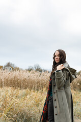 Young woman wearing grey old fashioned coat and tartan scarf stands in the dry grass reeds field looking at the camera.