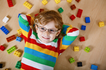 Little blond child with glasses playing with lots of colorful plastic blocks indoor. Kid boy wearing colorful shirt and having fun with building and creating