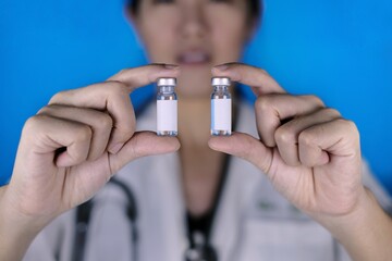 A female doctor holding 2 vials of vaccine in parallel. A physician in white coat and stethoscope...