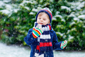 Cute little funny child in colorful winter fashion clothes having fun and playing with snow, outdoors during snowfall. Active outdoors leisure with children. Kid boy and toddler catching snowflakes.