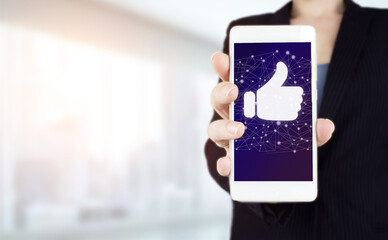Congratulate, good and like concept. Hand hold white smartphone with digital hologram hand tumb up sign on light blurred background. Tumb up concept.