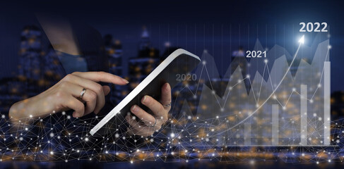 Happy New Year 2022 - Hand touch white tablet with digital hologram growth graph chart sign on city dark blurred background. Financial charts showing growing revenue In 2022