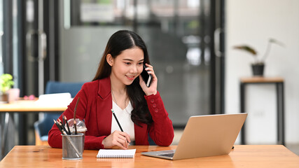 Obraz premium Smiling businesswoman checking information on laptop computer and talking on mobile phone.