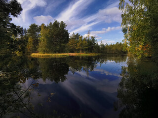 The mirror surface of a forest lake, in which trees and the sky with beautiful clouds are reflected.