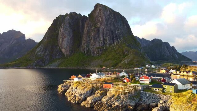 Drone flying over small fishing village Hamn&oslash;y in Norway towards iconic mountains above the sea with wooden drying racks for torrfisk
