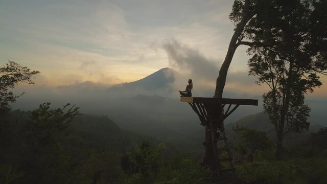 Woman in easy lotus pose on view platform with moody sunset at mount Agung, Bali