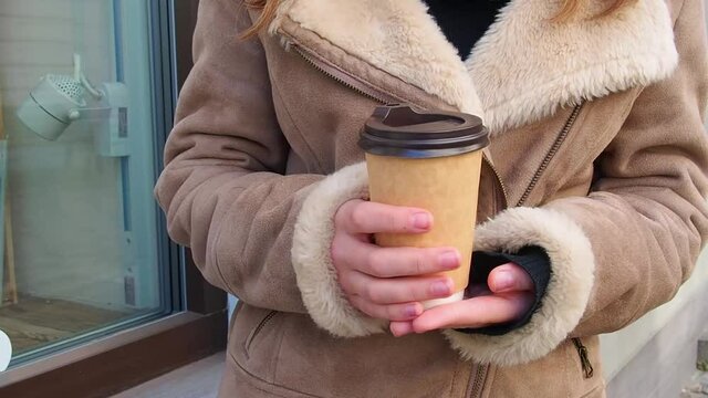 A young girl with blond hair in a brown coat walks down the street with takeaway coffee in cold winter day. Close up footage with hands holding paper cup with coffee. 