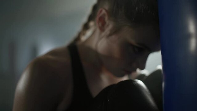 Woman power, tired female fighter stands pressed against a punching bag, hard training in the boxing gym, 4k slow motion.