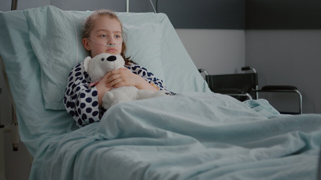 Alone worried child patient wearing oxygen nasal tube resting in bed holding teedy bear in hands recovering disease consultation in hospital ward. Lonely kid suffering fibrosis infection