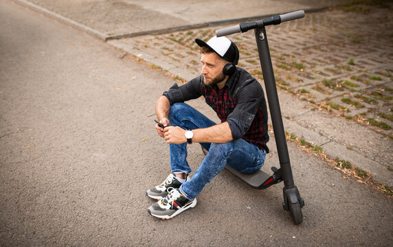 Top above high angle view photo of young urban man sitting on electrical scooter on the street listening to music wearing a hatholding his phone