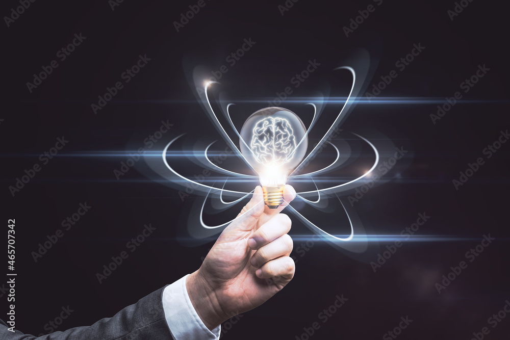 Poster close up of hand holding abstract light bulb on dark wide background with digital brain and atom out