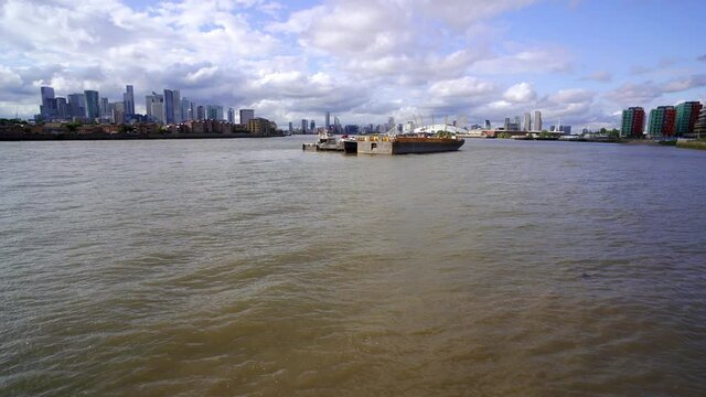 Tilting view revealing East London with Docklands and Canary Wharf on one side and Greenwich and the Millennium Dome on the other. Barges float in the middle of the wide River Thames