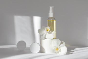 Fototapeta na wymiar Mock-up of glass bottle with essential oil, two white jars with cream, surrounded by orchid flowers. White background with morning rays of sun. Concept of skin care, spa treatments