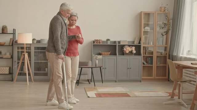Full shot of older Caucasian man with cane walking in living room at daytime, his caring Asian wife assisting him