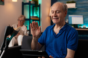 Fototapeta premium Retired adult waving at tablet with video call conference on sofa with crutches. Old man using online remote communication while elder disabled woman sitting in wheelchair reading