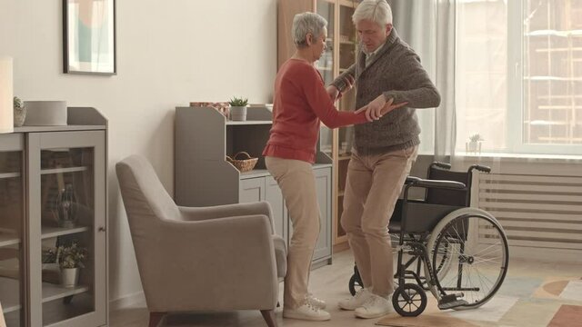 Full side view of caring short-haired Asian woman helping senior Caucasian man getting up from wheelchair and sitting down on armchair in living room at daytime