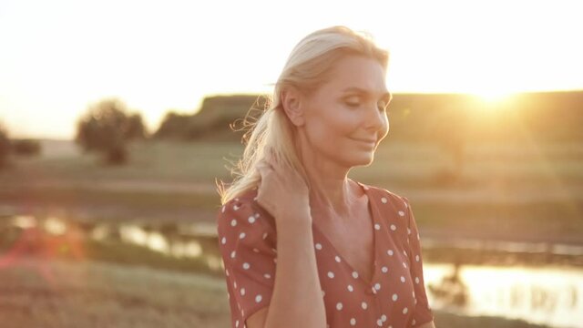 Happy woman correcting her hairstyle and walking on the field near the river