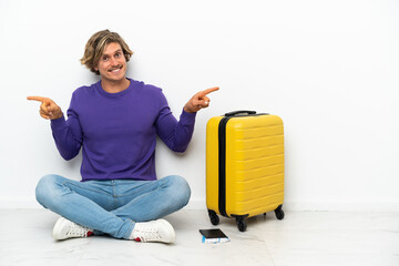 Young blonde man with suitcase sitting on the floor pointing finger to the laterals and happy