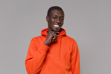 Smiling young african american man guy in orange streetwear hoodie posing isolated on grey background studio portrait. People emotions lifestyle concept. Mock up copy space. Put hand prop up on chin.