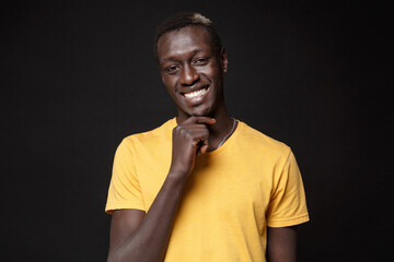 Smiling young african american man guy in yellow t-shirt posing isolated on black background studio portrait. People sincere emotions, lifestyle concept. Mock up copy space. Put hand prop up on chin.