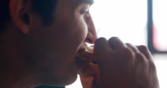 Close up shot of handsome men are eating burger in fast food restaurant. People eat unhealthy meal. Getting satisfaction from unhealthy food. Junk food, modern life concet. Side view, slow motion