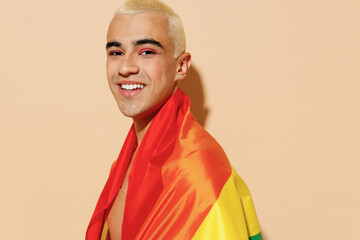Side view smiling cool young blond latin gay man 20s with make up in beige tank shirt wrapped in rainbow flag look camera isolated on plain light ocher background studio People lgbt lifestyle concept.