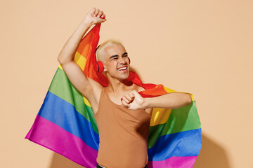 Young cool blond latin gay man with make up in beige tank shirt headphones wrapped in rainbow flag...